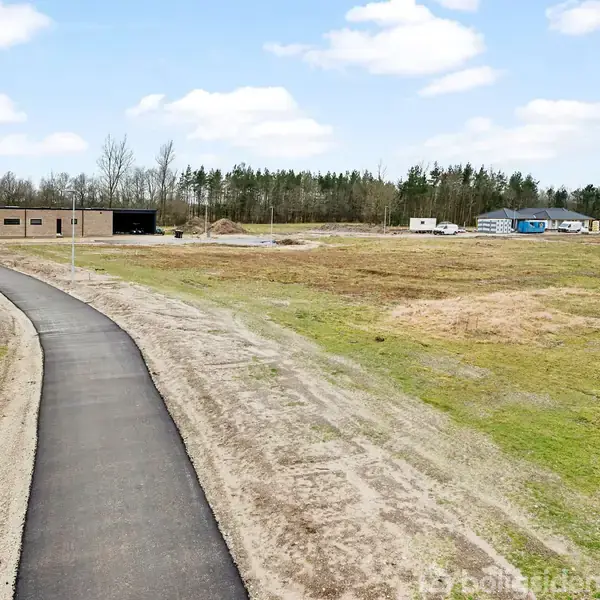 En asfalteret sti fører til en lysebrun bygning i et åbent, græs- og jorddækket landskab. Bygninger og nogle køretøjer ses i baggrunden, med en skovgrænse under en skyet himmel.