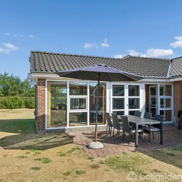 Rød murstensbolig med terrasse, hvor et sort bord og seks stole står under en parasol. Der er en grill ved væggen. Græsplæne og træer i baggrunden under blå himmel.