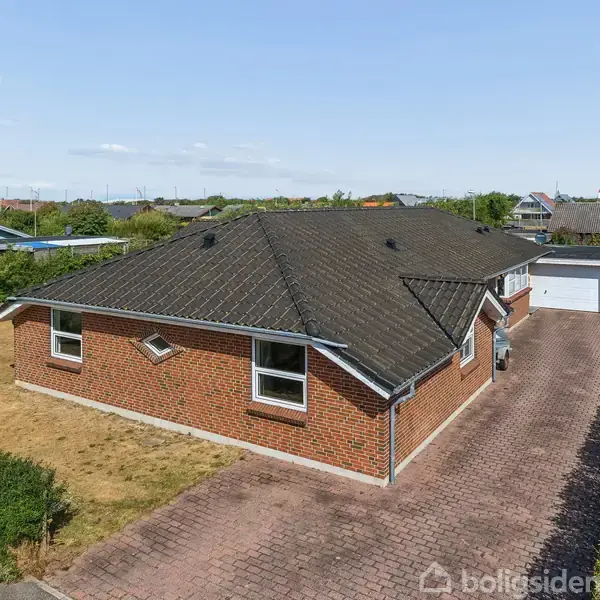 En murstensvilla står stille, omgivet af en veltrimmet have. Ved siden af er der en dobbeltgarage. Et dansk flag vajer i haven, og der er en indkørsel af belægningssten.