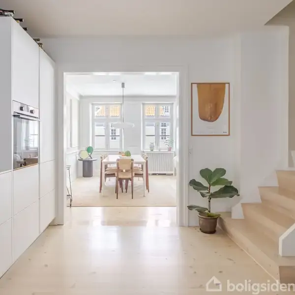 En moderne og lys spisestue med træbord og stole er synlig fra et køkken med hvide skabe. Trappe til højre. Plante og kunstplakat ved siden af.