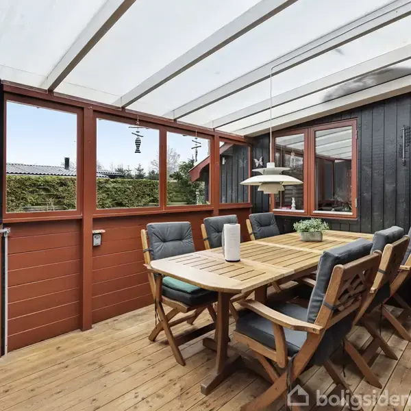 Et udendørs spisebord med seks stole står på en træterrasse. Rummet er overdækket med glas og omgivet af trævægge og et hegn af buske i baggrunden.