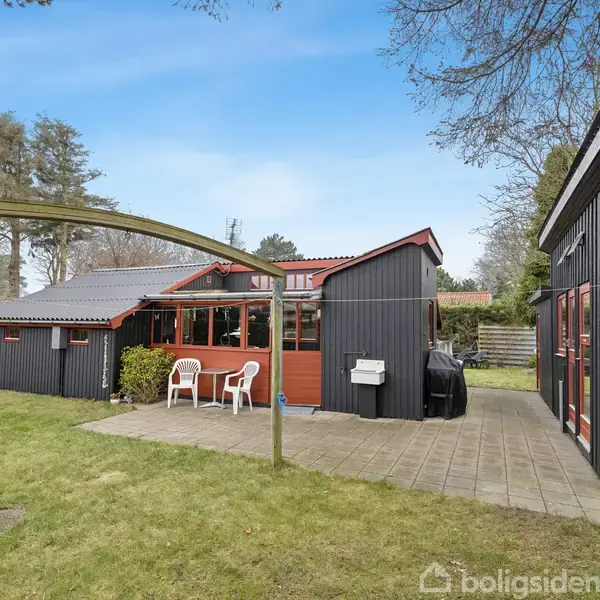 En sort trælængebygning med røde detaljer står i en grøn have. To hvide plaststole foran huset, og flisebelagt terrasse. Træer og hegn omringer området.