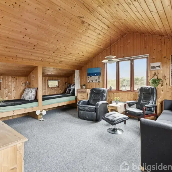 En hyggelig stue med trævægge og -loft. To lænestole med en fodskammel står foran vinduerne. En sofa og seng er placeret i rummet, med planter som dekoration.