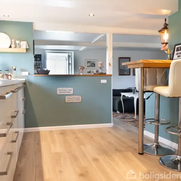 Køkken med hvide skabsfronter og træbordplade. Høj stol ved bardisk. Grønne vægge dekoreret med hylder. Tekst på væggen: "Mum's Kitchen Rules" og "P.S. Don't forget to smile."