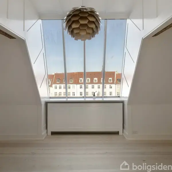 Et opholdsrum med en stor, skrå loftsvindue, der viser en udsigt over bygninger med røde tage. En radiator er placeret under vinduet. Loftlampe hænger ned fra loftet.