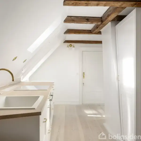 Køkken med hvid bordplade og vask, gylden vandhane i et skråloft rum. Synlige træbjælker i loftet og en dør for enden af rummet. Veggene er hvide.