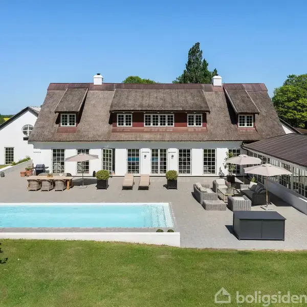 Hus med stråtag og mange vinduer, har en pool og terrasse med havemøbler. Det ligger i en landskabelig, grøn have med marker og træer i baggrunden. Ingen tekst findes.