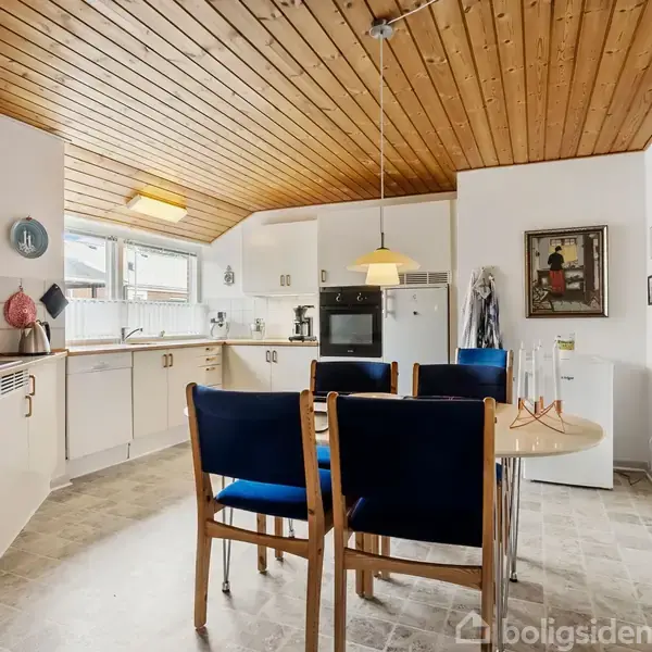 Et køkken med hvidt inventar og træloft indeholder et rundt bord med blå stole omkring. Baggrunden viser en ovn og billedramme på væggen. Ingen tekst er fundet.