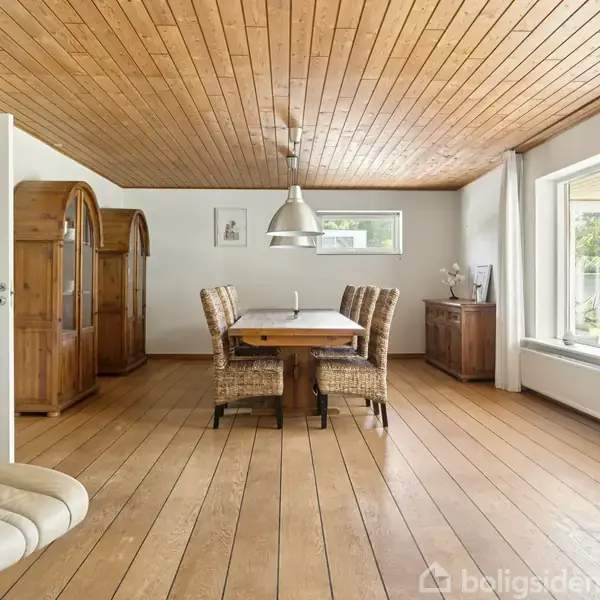 Et spisebord med stole står i et rum med trægulv og loft. Et stort vindue til højre lukker naturligt lys ind, mens en kommode og vitrineskab er i baggrunden.