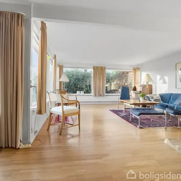 En stue med trægulv indeholder en blå sofa og et sofabord på et tæppe. Store vinduer lukker lys ind, og udvendigt ses et grønt landskab.