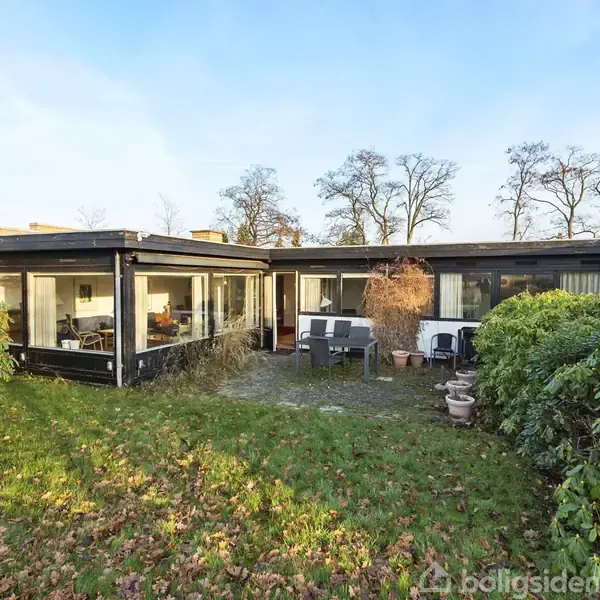 En etplansvilla står stille i en have med græsplæne og omkranset af buske. Store vinduer afslører en spisestue indenfor. Udendørs terrassemøbler ses foran huset under åben himmel.