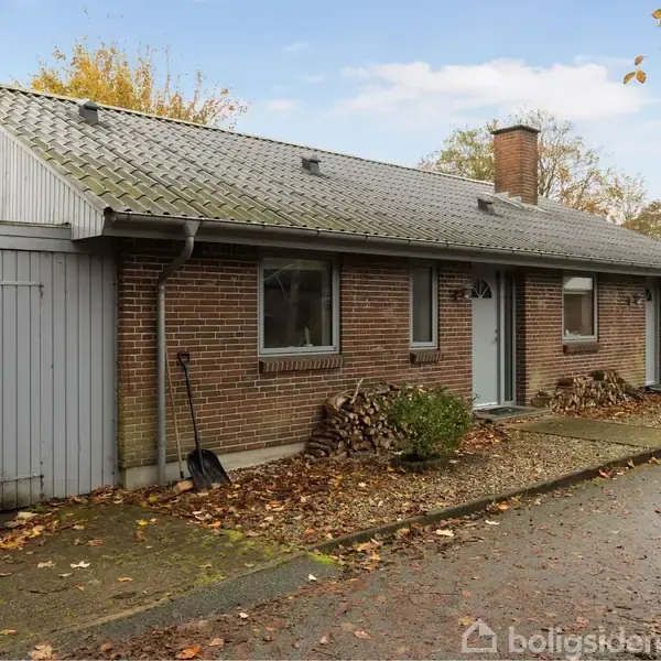 En murstenshus står stille ved en vej, omgivet af efterårsblade og spredte buske. Et skur til venstre, en stabel brænde foran huset, og en grå himmel i baggrunden.