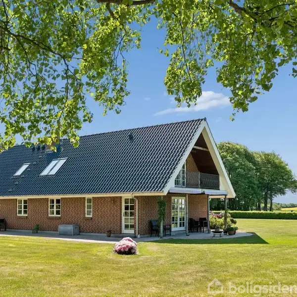 Et rødt murstenshus med sort tag ligger i en frodig have. Huset har flere vinduer og en lille overdækket terrasse. Store træer grænser op til græsplænen under en blå himmel.