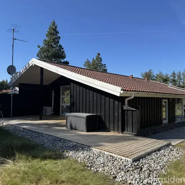 Et sort sommerhus med rødbrunt tag står på en stenbelagt terrasse i en skovklædt område. Der er en satellitantenne og en stor vinduesfacade med omgivelser af grønne træer.