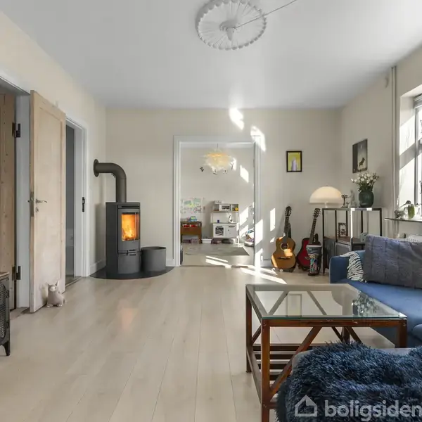 Stue med blå sofa ved et stort vindue. En brændeovn i hjørnet afgiver varme. Et sofabord og en lampe ved siden af sofaen. En guitar står ved væggen.