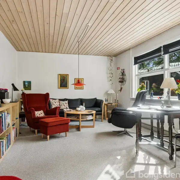 Rummeligt lyst stueområde med rødt lænestolssæt, rundt sofabord, sort sofa og sort kontorstol ved et træbord. Hylder, billeder, og store vinduer med udsigt til have.