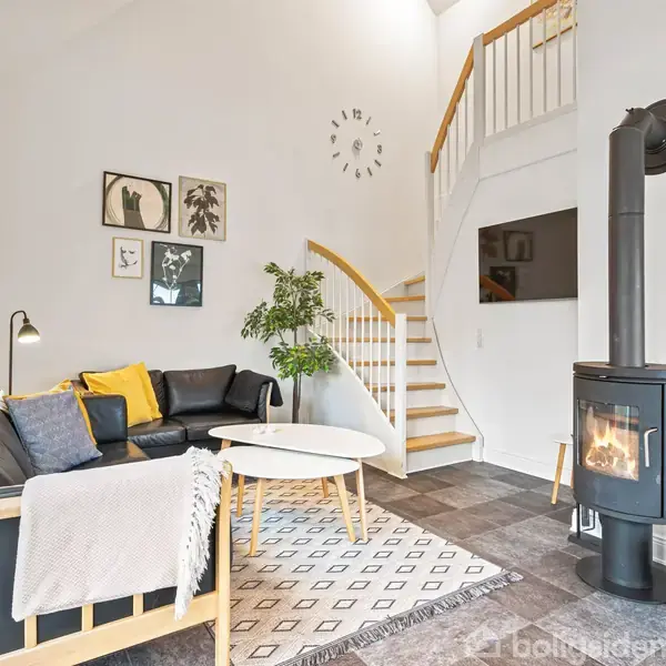 I en lys stue står en sort sofa med gule puder ved siden af et rundt bord. En brændeovn er tændt. Trappe fører opad. Vægur viser klokken.