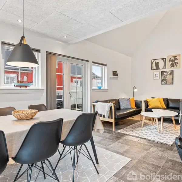 Et moderne spisebord med sorte stole står i et lyst rum, del af en stue med sofa, billeder på væggen og en brændeovn.