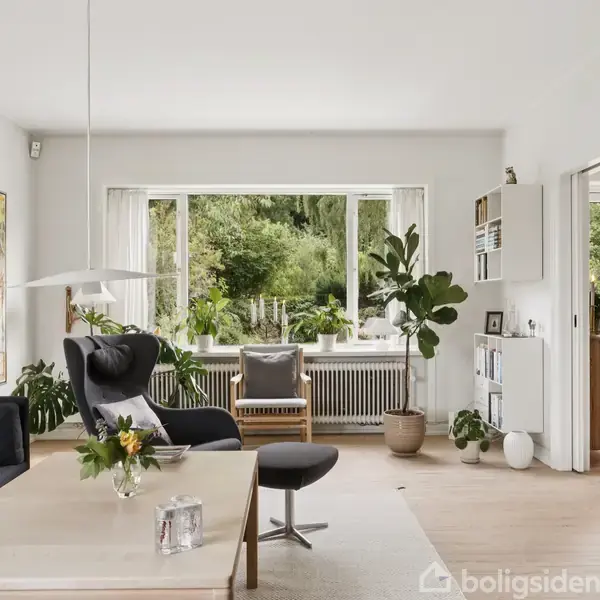 Stue med sofa og stole arrangeret omkring et sofabord, omgivet af store vinduer med udsigt til en grøn have. Plakater og planter dekorerer rummet, der er lyst og luftigt.