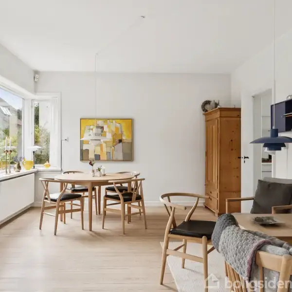 Stue med lyse trægulve, hvor et træbord og stole er centralt placeret. Væggene har moderne kunstværker. Til højre ses en høj skænk og en blå lampe.