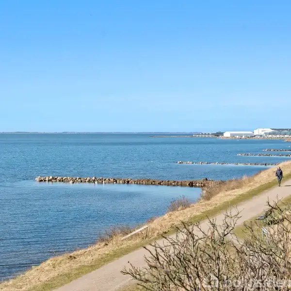 Sti ved vandet med en person, der går langs en grusvej. Havet strækker sig mod horisonten, mens buske omgiver stien. Bygninger er synlige i baggrunden.