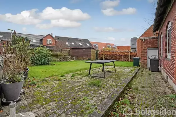 En tom havebord står på en flisebelagt terrasse ved siden af et rødt murstenshus. Græsplænen grænser op til hegn og nabohuse i baggrunden.