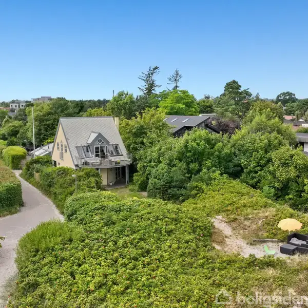 Et hus med tagterrasse står omgivet af frodig vegetation og stier. Omkringliggende huse er synlige i den grønne, landlige baggrund under en klar blå himmel.