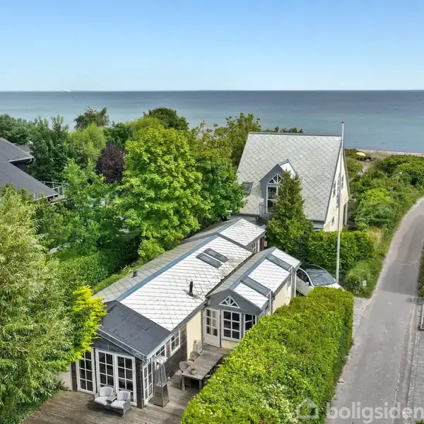 Et hus med lysegråt tag ligger omgivet af grønne buske og træer. Stien ved siden af fører til en strand med roligt hav i baggrunden under en klar blå himmel.