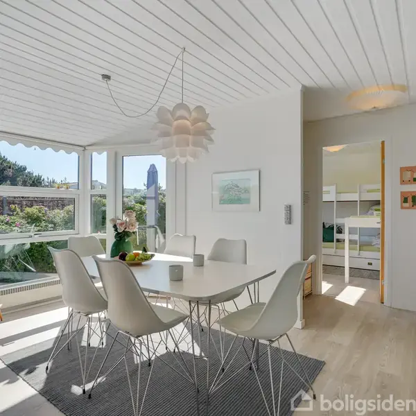 Spisebord står med seks stole omkring, i en lys og moderne spisestue med store vinduer. En lampe hænger over bordet, omgivet af et lyst og luftigt rum med udsigt til grønne områder.