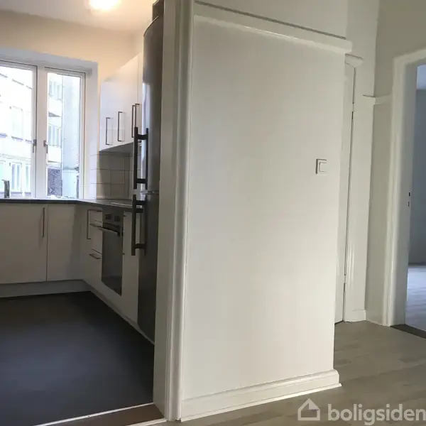 Et moderne køkken med hvide skabe og en indbygget ovn står venstre for en hvid væg. Et vindue giver udsigt til en bygning udenfor.