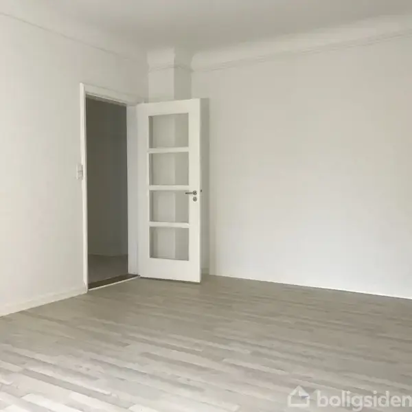 Dør står åben i et lyst rum med hvide vægge og lyst trægulv, hvilket giver en ren, minimalistisk følelse.