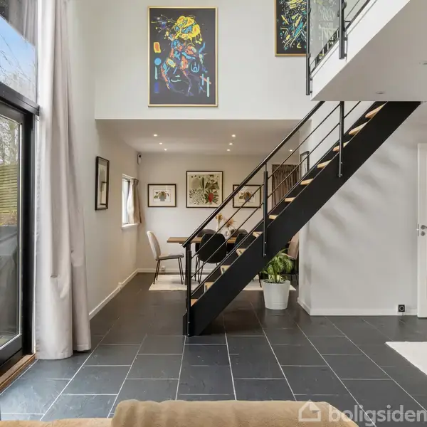 Stue med sort ståltrappe, der fører op til en åben etage. Store vinduer giver naturligt lys, og kunstværker hænger på væggene. Under trappen står en plante.