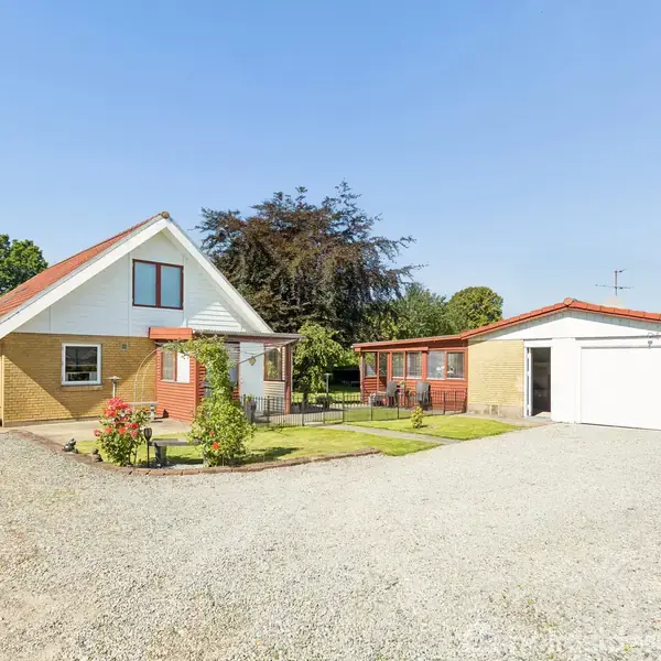 Et gult murstenshus med rødt tag står ved siden af en fritstående garage. Huset omkranses af en grusindkørsel og velholdt have i et solrigt villakvarter.