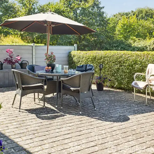 En udendørs terrasse med et rundt bord og fire stole under en stor parasol. Bordet har frugt og drikkevarer. Omkring er der hække og en lav mur med blomster.