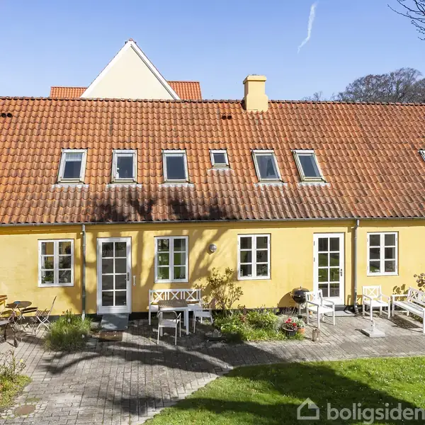Gul facade på rækkehus med rødt tegltag og flere vinduer. Havemøbler står placeret på flisebelagt terrasse med grøn græsplæne foran. Klar blå himmel i baggrunden.