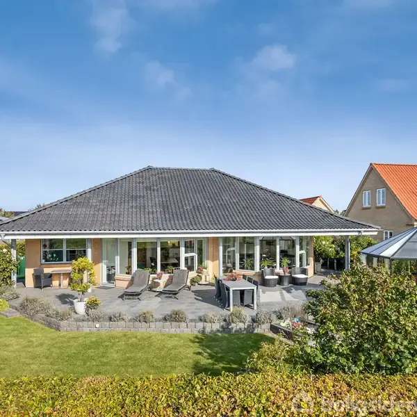 Hus med sort tag står i en velplejet have. Terrassen er fyldt med havemøbler under en blå himmel. Til højre ses en udestue af glas.