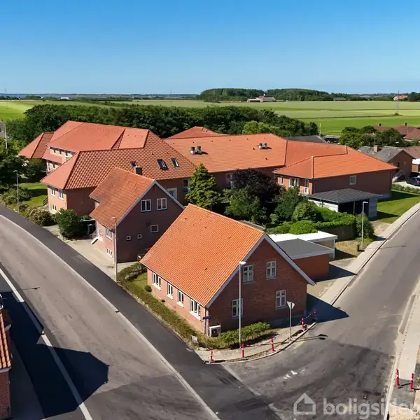 Røde murstensbygninger med orange tegltage står langs en vej i en lille by. Vejene danner en Y-krydsning, og området er omgivet af marker og træer under en klar blå himmel.
