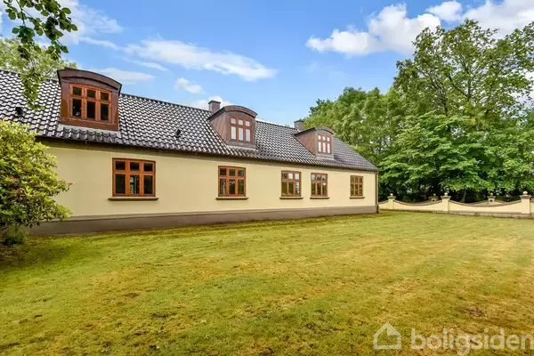 Et gult hus med sort tegltag og flere kviste står i en have med græsplæne. Der er træer og en hvid mur i baggrunden under en blå himmel.