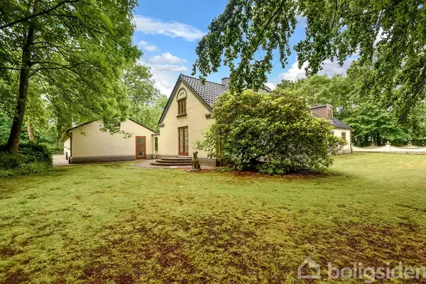 Et gult hus med spidst tag står omgivet af en frodig have. Træer og buske omgiver huset, og græsplænen strækker sig langt omkring. Himlen er let overskyet.