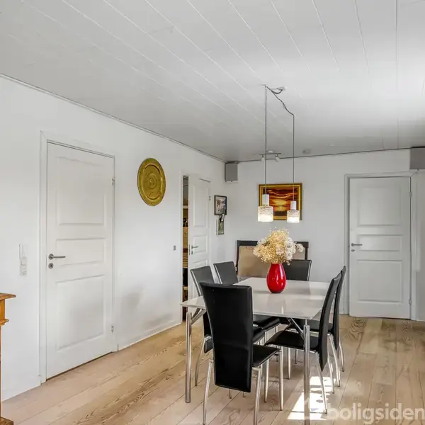 Spisebord med sorte stole står i et lyst rum med hvide vægge og trægulv. En rød vase med blomster pryder bordet, omgivet af døre og dekorative elementer.