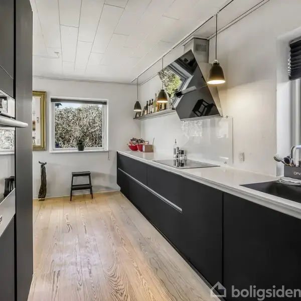 Et moderne køkken med sort og hvidt design. Det har en indbygget ovn, kogeplade, håndvask og vindue med udsigt til en have. Trægulv og hylder til dekoration.