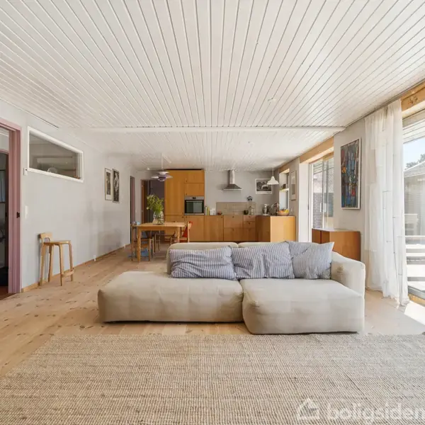Sofa står i en lys stue med trægulv, vendt mod et åbent køkken. Rummet har store vinduer med gardiner, der giver udsigt til terrasse.
