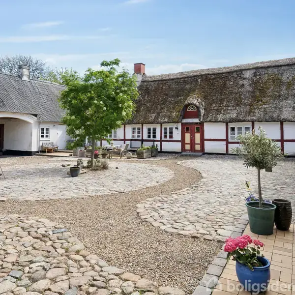 Gårdsplads i centrum med brosten, omgivet af bindingsværkhuse med stråtag. En busk og flere potteplanter står i gården, skaber en landlig og fredelig atmosfære.