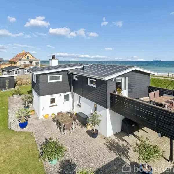 Hus med terrasse ligger ved kysten. Det sorte og hvide hus har en stor, møbleret balkon. Græsplæne og havet danner en naturskøn baggrund.