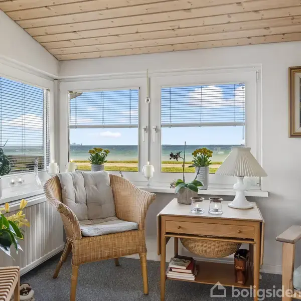 Kurvestol med hynde står foran vindue med udsigt til havet. Bord med lampe og planter ved siden af. Blå himmel og skyer udenfor.
