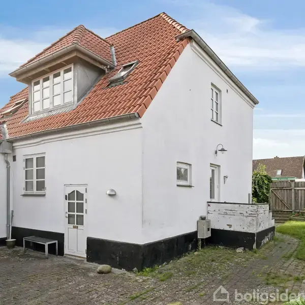 Hvidt hus med rødt tegltag, ingen synlig handling, beliggende i en gård med flisebelægning og græsplæne. En parasol læner sig mod en mur, og et plankeværk omkranser.