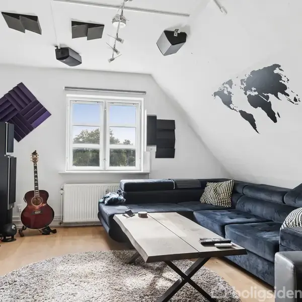 En stue med en mørk sofa omkring et træbord. En guitar står op ad en reol ved siden af et vindue. En verdenskort-vægdekoration hænger på skråvæggen.