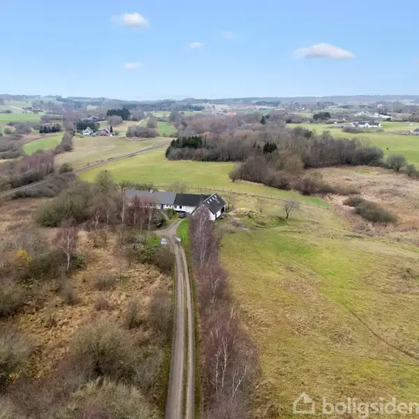 Et landhus ligger isoleret langs en snoet vej, omgivet af åbne marker og let bevoksede områder under en klar blå himmel med enkelte skyer.