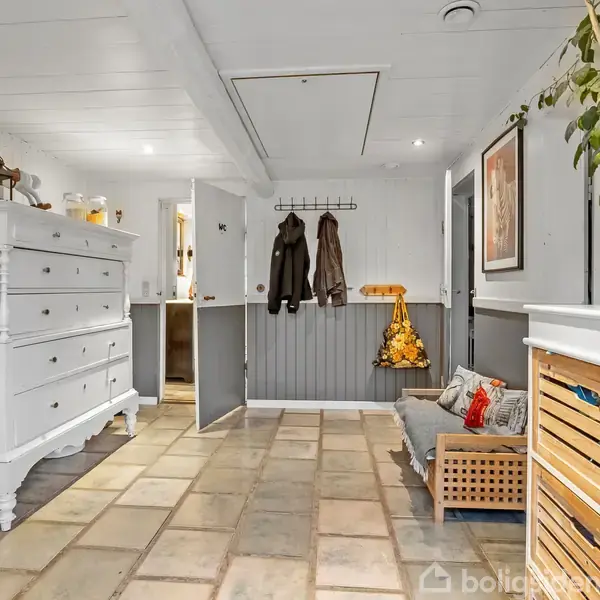 Kommode er placeret i en korridor med lyse fliser, omgivet af en bøjle med jakker, en sofa med puder og planter. Rummet har træpaneler og vægdekorationer.