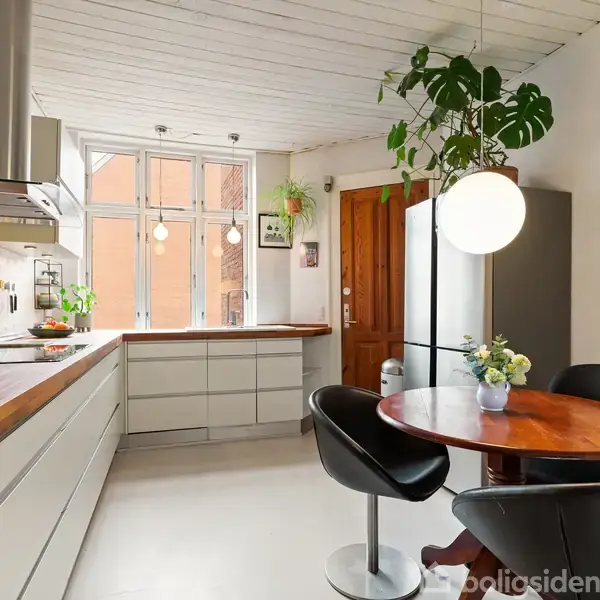Et køkken viser moderne hvide skabe og en træbordplade. Der hænger en stor lampe over et rundt træbord med sorte stole. Vinduerne lader naturligt lys ind, og planter pynter rummet.